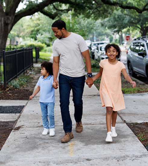 A parent walks down a residential sidewalk holding the hands of their two young children. 