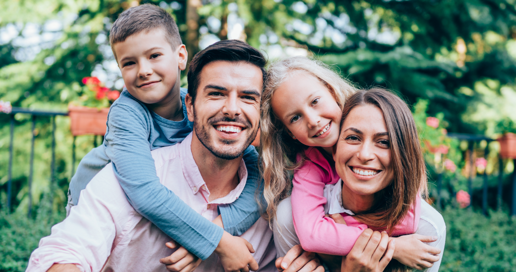A family of two adults and two children smile at the camera while embracing outdoors.